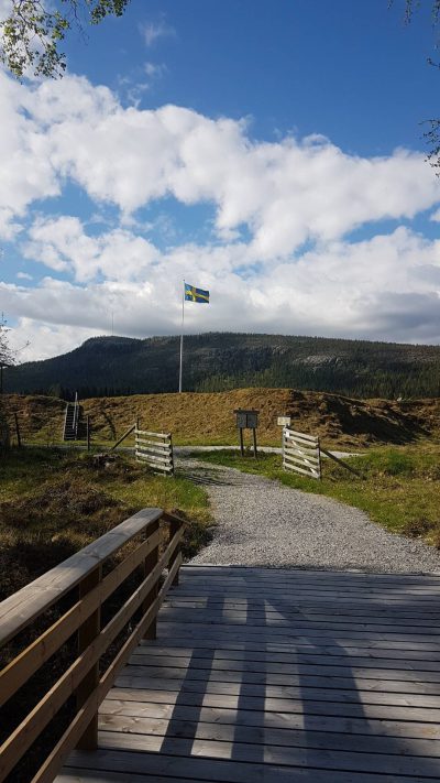 Svenska flaggan hissad inne på Långå Skans. I förgrunden ses bron över Vallgravsbäcken och jordvallarna runt skansen. Foto: Liselott Prestjan, Projektgruppen för Långå Skans